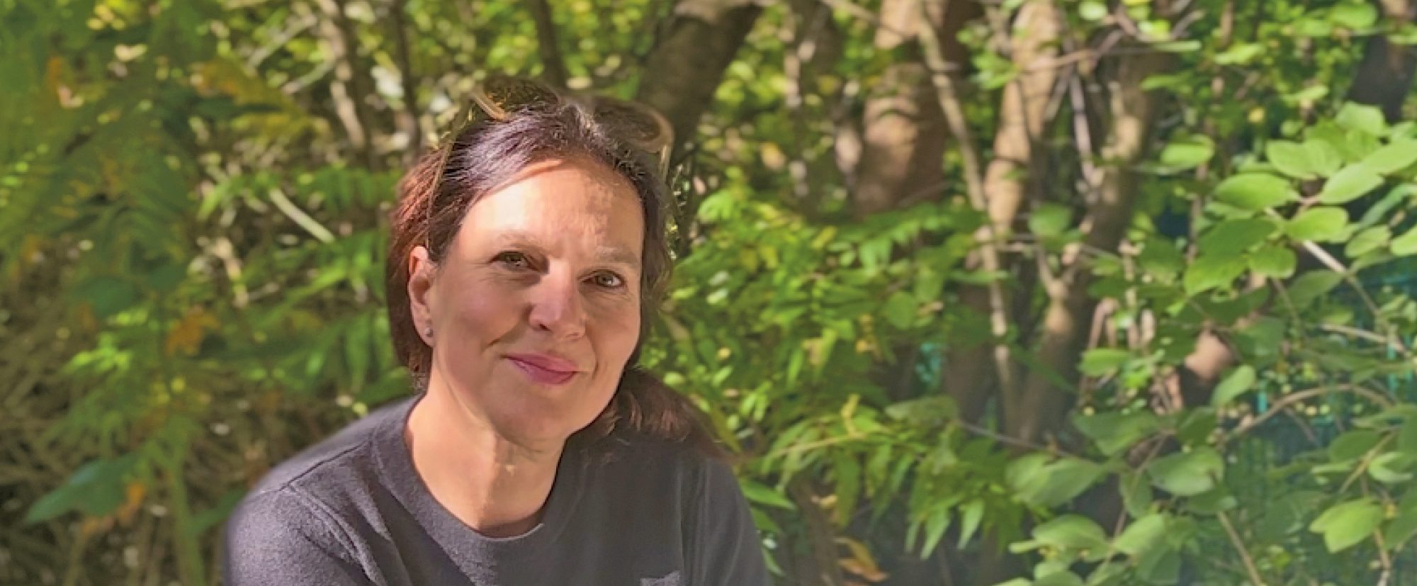 Charlotta Móna sitzt im Garten, hält ein Buch in der Hand und lächelt in die Kamera einladend