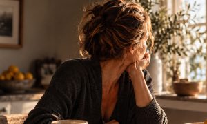 Nachdenkliche Frau an einem sonnigen Tisch mit Kaffee, Smartphone und Notizbuch.
